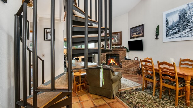A living room with a fireplace, a dining area with wooden chairs, and a staircase with metal railings.
