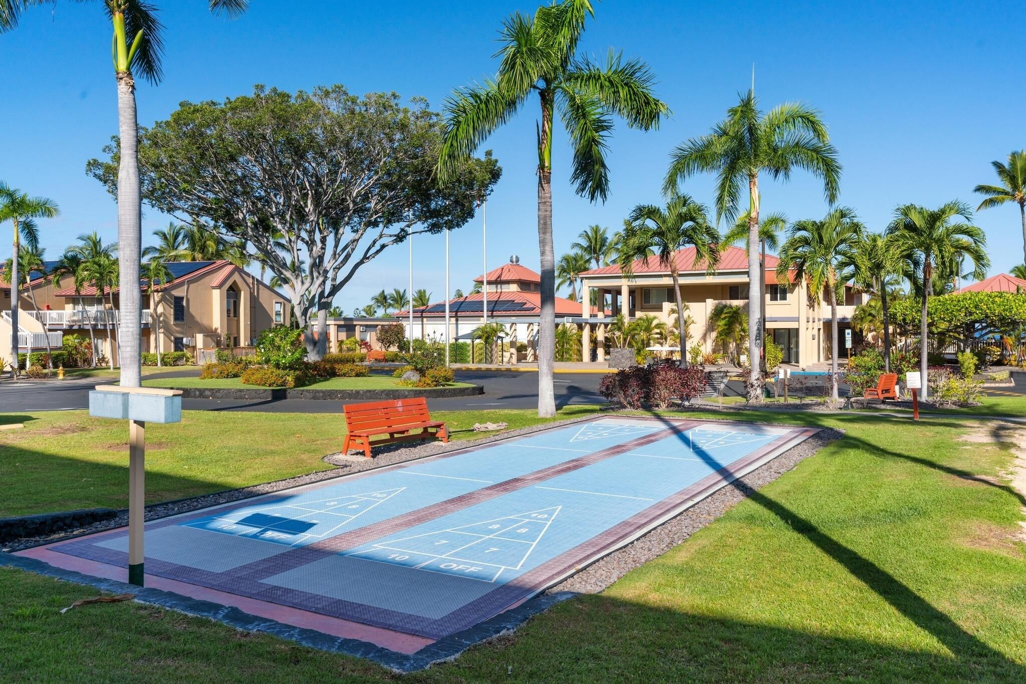 Un patio con cancha de shuffleboard, palmeras y un edificio de techo rojo.