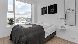 A modern bedroom with a large window, a bed with a dark gray bedspread, two wall-mounted artworks, a bedside table with a lamp, and a mirror. - Spacious 3BR apartment with 2 bathrooms (Reykjavík)