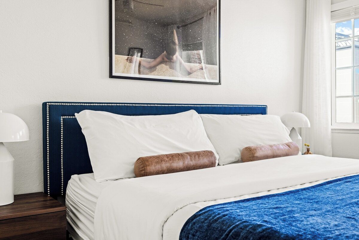 A neatly made bed with white linens and a blue blanket, a framed picture on the wall, and a bedside lamp.