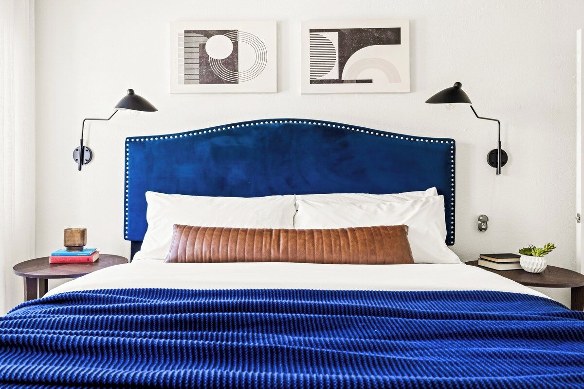A bed with a blue headboard, white and brown pillows, and a blue textured blanket. There are two wall-mounted lamps on either side of the bed.