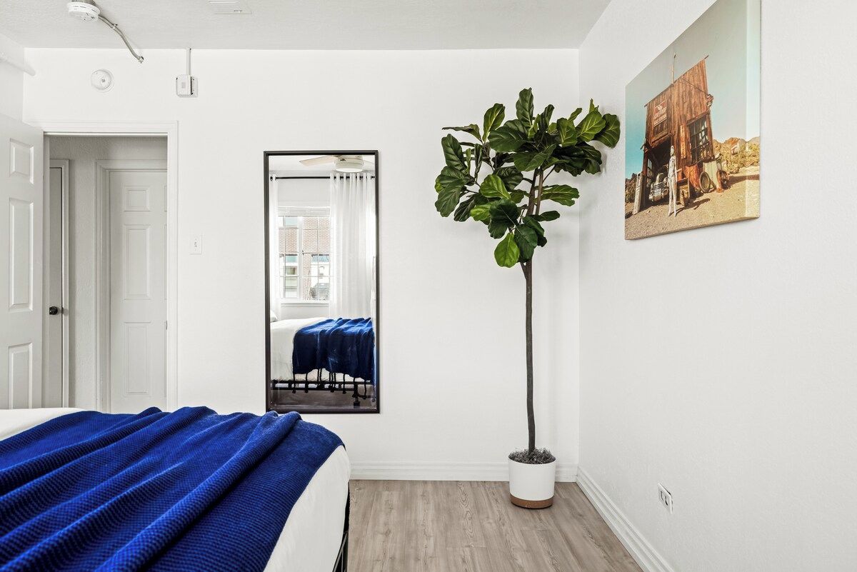Un dormitorio con cama, espejo, planta en maceta y un cuadro en la pared.