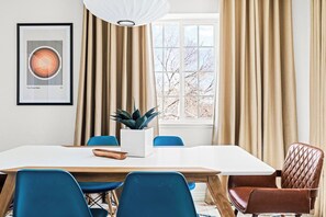 Un comedor moderno con una mesa blanca, sillas azules y una butaca de cuero marrón. Un cuadro enmarcado decora la pared, y hay una planta sobre la mesa.