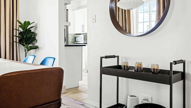 Habitación de hotel moderna con un espejo redondo, una mesita con vasos, una planta en maceta y vistas a la cocina y la sala de estar.