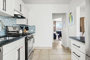 Una cocina con armarios blancos, salpicadero de azulejos y vistas a un salón con sofá y mesa.