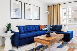 A living room with a blue sofa, a white side table, a wooden coffee table, and framed artwork on the wall.
