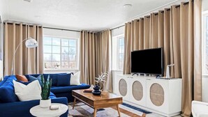 Una sala moderna con un sofá azul, una mesa de centro de madera, una mesita auxiliar blanca, un televisor de pantalla plana sobre un mueble blanco y cortinas color beige.