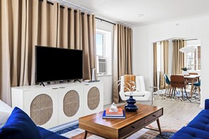 Un salon moderne doté d’un téléviseur à écran plat, de meubles blancs, d’une table basse avec des livres et d’un coin repas comprenant des chaises et une table.