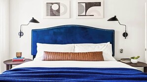 Una cama bien hecha con cabecera azul, dos lámparas de noche y una planta pequeña en la mesita.