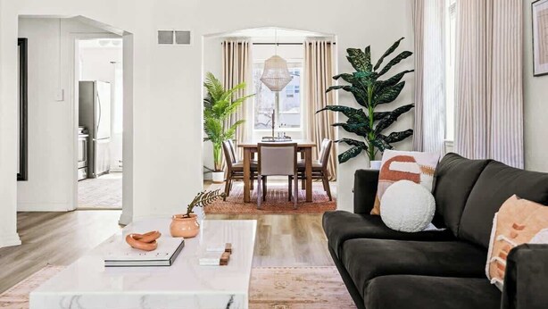 Een moderne woonkamer met een donkere bank, een witte salontafel en een eethoek met een tafel en stoelen.