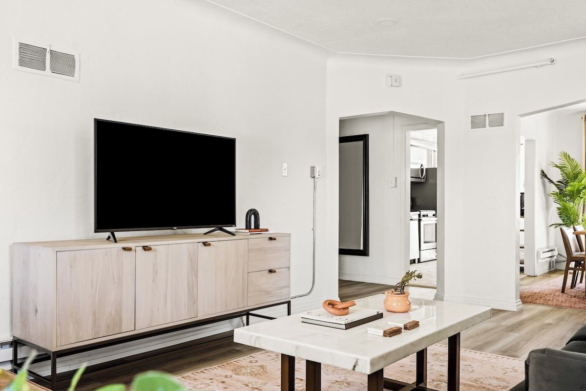 Een moderne woonkamer met een flatscreen televisie, een houten kast, een marmeren salontafel en uitzicht op de keuken.
