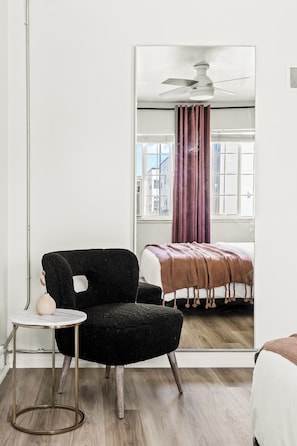A room with a black armchair, a small round table, a bed with a brown blanket, and a window with curtains. - Modern Comfort | King Beds | Sloans Lake | 30 Day (Denver)