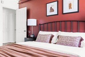 Une chambre à coucher avec un lit, des murs blancs, un mur d’accent rouge et des œuvres d’art encadrées.