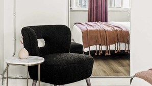 Een moderne kamer met een zwarte fauteuil, een klein rond tafeltje en een bed met een bruine deken.