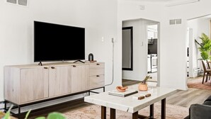 Een moderne woonkamer met een flatscreen televisie bevestigd aan een licht houten kast, een witte marmeren salontafel met decoratieve objecten, en een uitzicht op de keuken.