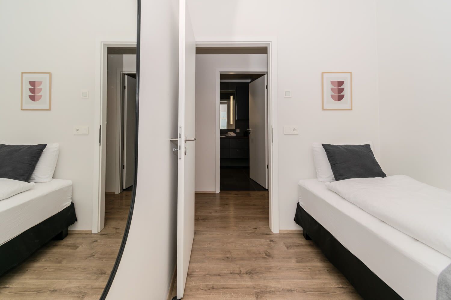A room with two single beds, a wooden floor, and a hallway leading to a bathroom.