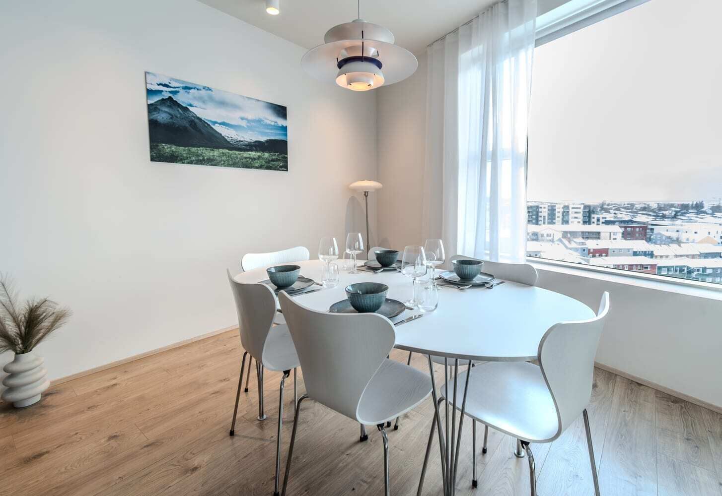 A modern dining room with a round table, white chairs, and a city view through large windows.