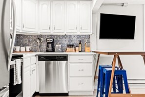 A modern kitchen with white cabinets, a dishwasher, a coffee maker, and a flat-screen TV mounted on the wall.
