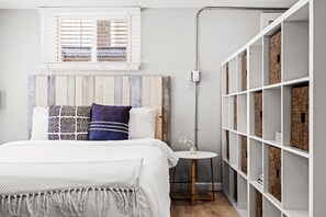 Un dormitorio con una cama, una mesita de noche, una ventana con persianas, un estante en la pared con cestas y un cabecero de madera.