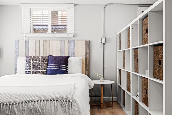 Une chambre à coucher comprenant un lit, une table de chevet, une fenêtre avec des stores, une étagère murale avec des paniers et une tête de lit en bois.