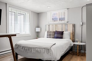 A bedroom with a bed, bedside table, lamp, and windows. - Contemporary Studio Near Sloan's Lake | 30 Day (Denver)