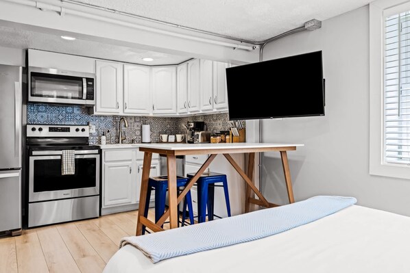 Una cocina funcional con armarios blancos, microondas, horno y lavaplatos. Hay una mesa de madera con taburetes azules y un televisor de pantalla plana montado en la pared.