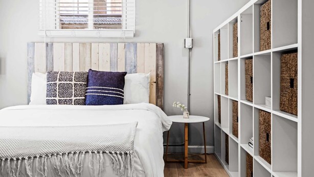 A bedroom with a bed, a bedside table, a window with blinds, a wall-mounted shelf with baskets, and a wooden headboard. - Contemporary Studio Near Sloan's Lake | 30 Day (Denver)