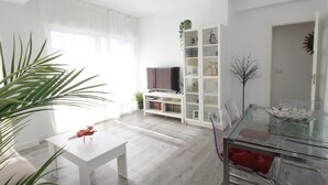 Un salón moderno con una mesa de comedor de vidrio, muebles blancos, un televisor sobre un mueble bajo y una estantería.