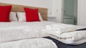 Una cama bien hecha con ropa blanca y almohadas rojas, una toalla blanca doblada sobre la cama, y una colcha de color oscuro.