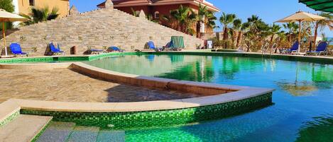 Una piscina con bordes de azulejos y sillas de descanso, rodeada de palmeras y un muro de piedra.