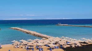 A beach with numerous sun umbrellas and lounge chairs, a rocky breakwater, and a clear blue sea.