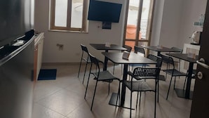 A dining area with tables and chairs, a flat-screen TV mounted on the wall, and a window with a view of the outside.