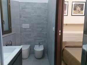 A bathroom with a bidet, a sink, and a toilet. There is a mirror on the left and a door leading to a bedroom. - Arte Mia b&b (Reggio Calabria)