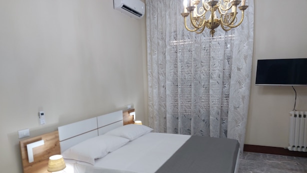 A hotel room with a bed, bedside lamp, air conditioning unit, chandelier, curtains, and a TV.
