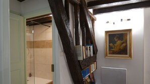 Une salle de bain avec une douche, un carrelage au sol, une poutre en bois et une bibliothèque contenant des livres, ainsi qu’un tableau encadré accroché au mur.