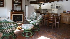 Un salon chaleureux avec une cheminée, des fauteuils en osier vert et un coin cuisine doté de meubles blancs et d’un four à micro-ondes.