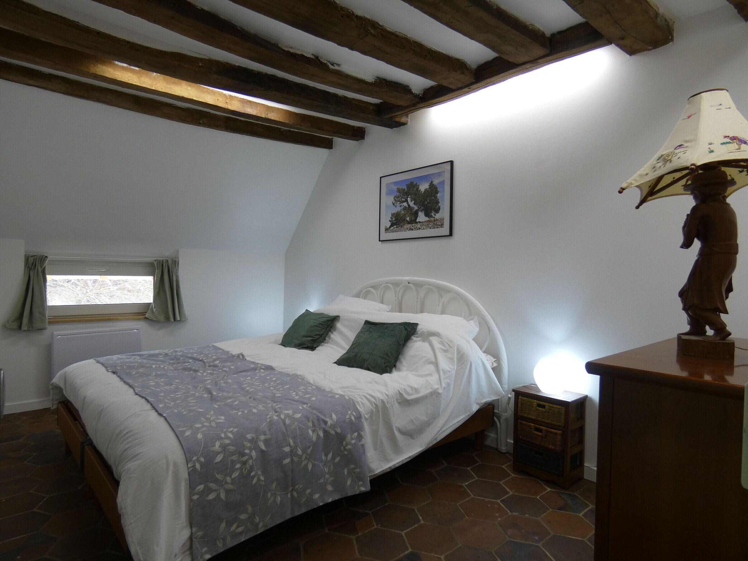 Une chambre à coucher comprenant un lit, une table de chevet, une lampe, une fenêtre avec des rideaux et un tableau accroché au mur.