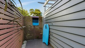 Un sentiero esterno stretto che conduce a un'asse di surf blu appoggiata a un muro, con una recinzione in legno da un lato e un cancello metallico dall'altro.