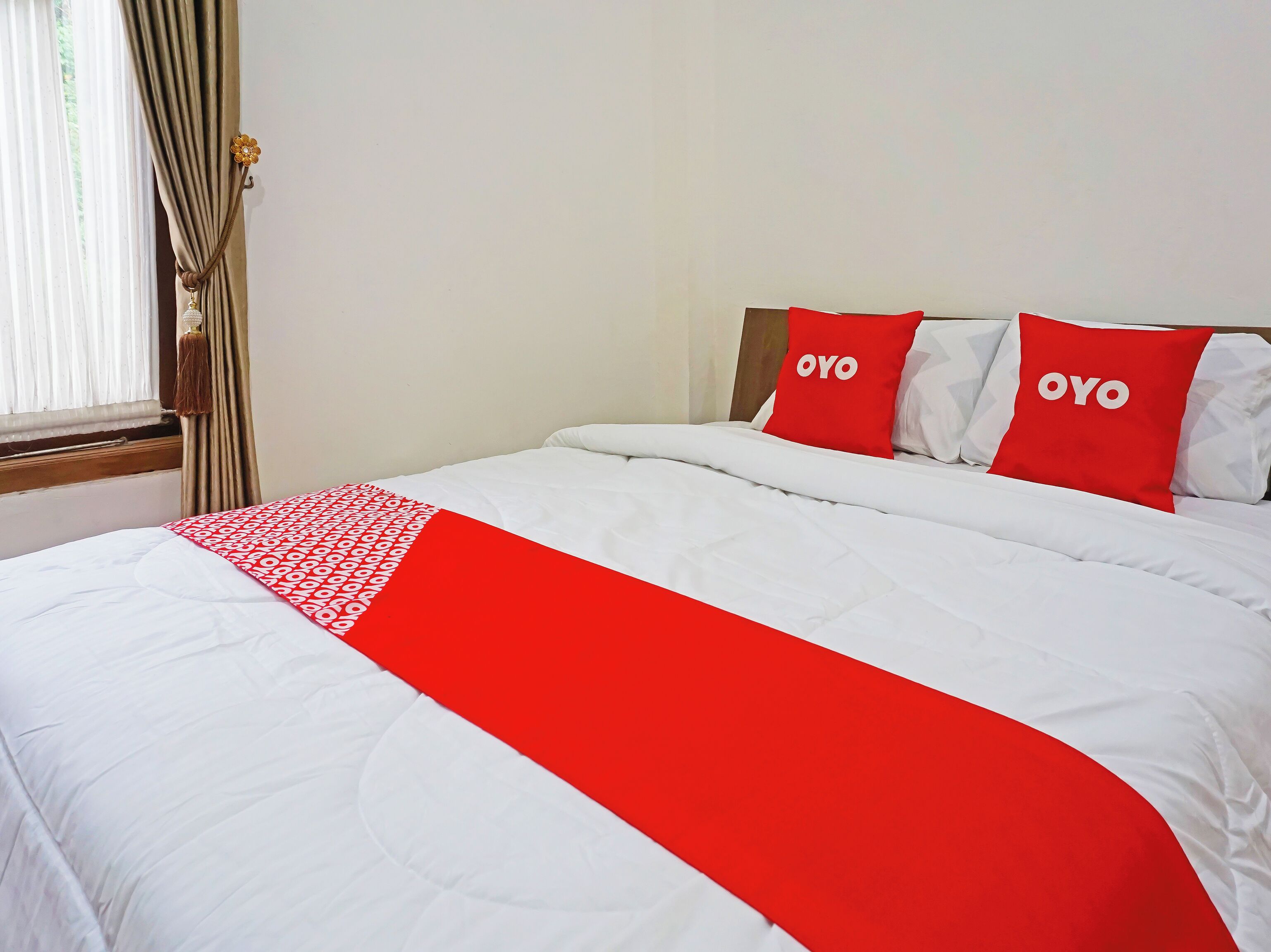 Una habitación de hotel con una cama, dos almohadas con la palabra 'OYO' y una colcha roja y blanca, además de una ventana con cortinas.