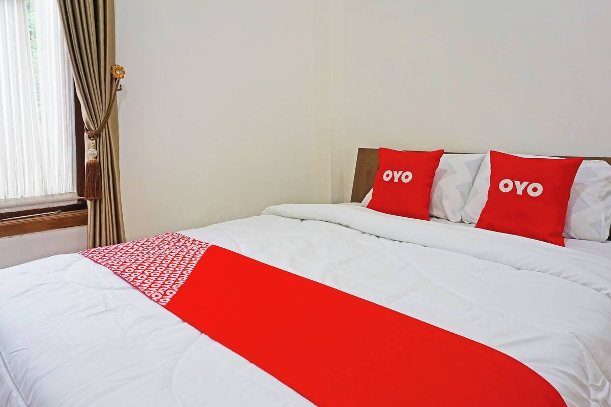 Een hotelkamer met een bed, twee kussens met de tekst 'OYO' erop, een rood-witte dekbedovertrek en een raam met gordijnen.