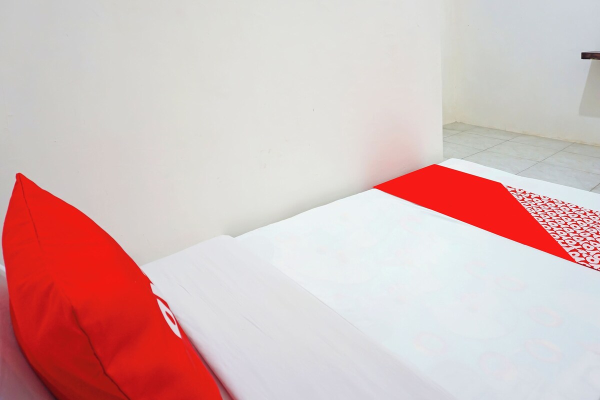 Een bed met witte beddengoed en rode accenten, een effen witte muur en een betegelde vloer.