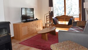 Een gezellige woonkamer met een televisie, houten meubels, een open haard en een groot raam met gordijnen.