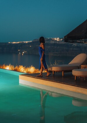 A person walking along a poolside area with lounge chairs and a view of a body of water and distant lights. - Divino Caldera (Santorini)