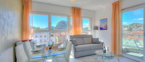A modern living room with a glass table, white chairs, a grey sofa, and large windows with curtains.