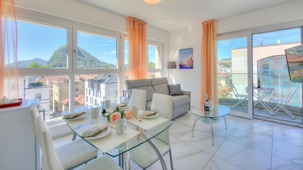 A modern living room with a glass dining table, white sofas, and a balcony with outdoor seating. - Attico al Parco: Penthouse in Lugano Balcony, A/C & 2 min From Lake (Lugano)