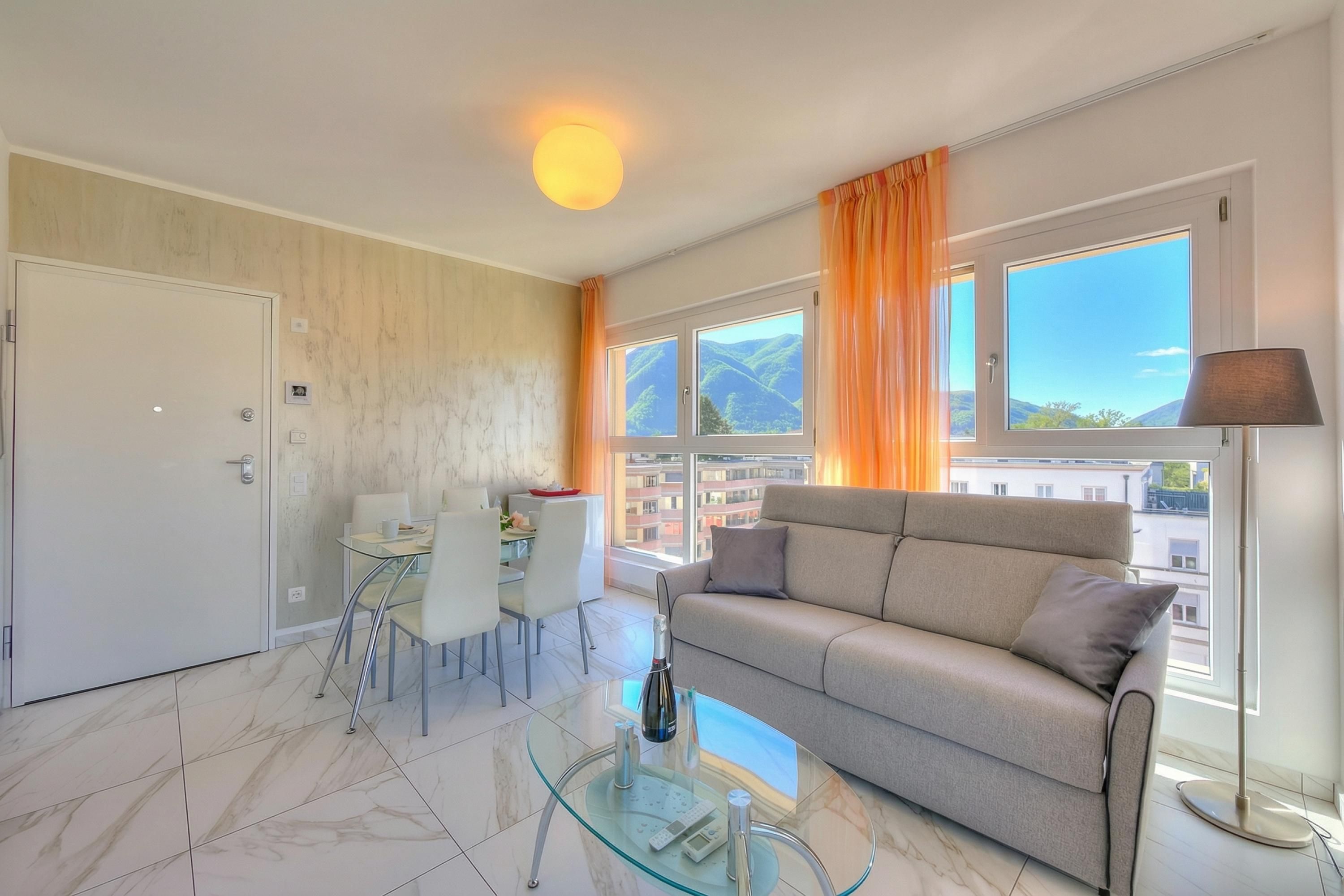 A modern living room with a sofa, a glass coffee table, a dining area with chairs, and large windows with curtains.