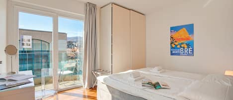 A hotel room with a bed, a desk, a chair, a wardrobe, and a balcony with a view of the city.