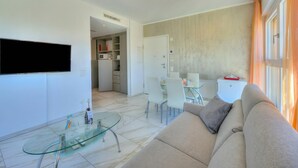 A modern living room with a glass coffee table, a beige sofa, a flat-screen TV mounted on the wall, and a dining area with white chairs and a glass table. - Attico al Parco: Penthouse in Lugano Balcony, A/C & 2 min From Lake (Lugano)