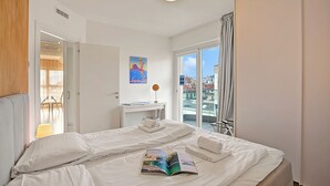 A hotel room with a bed, a reading area with a lamp, a window with curtains, and a view of buildings outside. - Attico al Parco: Penthouse in Lugano Balcony, A/C & 2 min From Lake (Lugano)