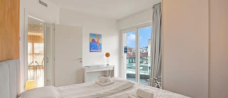 A hotel room with a bed, a reading area with a lamp, a window with curtains, and a view of buildings outside.